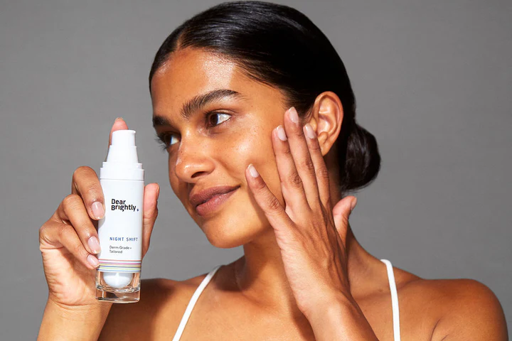 A person in a white top applies a skincare product to their face, holding a bottle labeled "Dear Brightly.