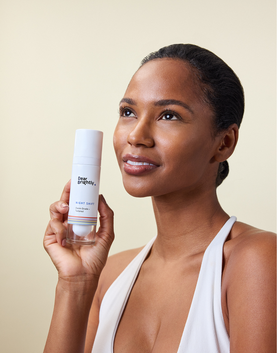 A woman in a white top holds up a bottle of skincare product labeled "Don't Brighten Me Up Night Shift" while looking upwards and smiling.