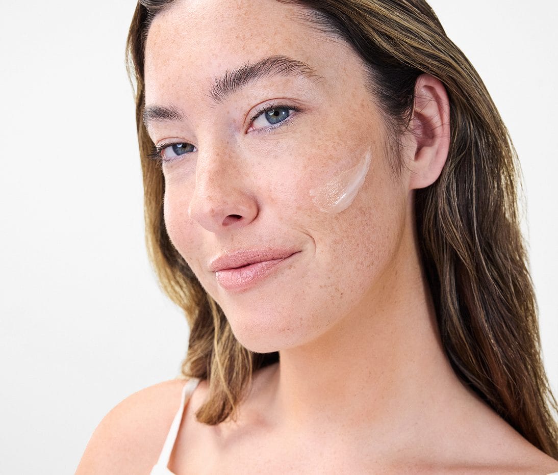 A woman with light skin and brown hair looks at the camera, wearing minimal makeup and a swipe of cream on her cheek.
